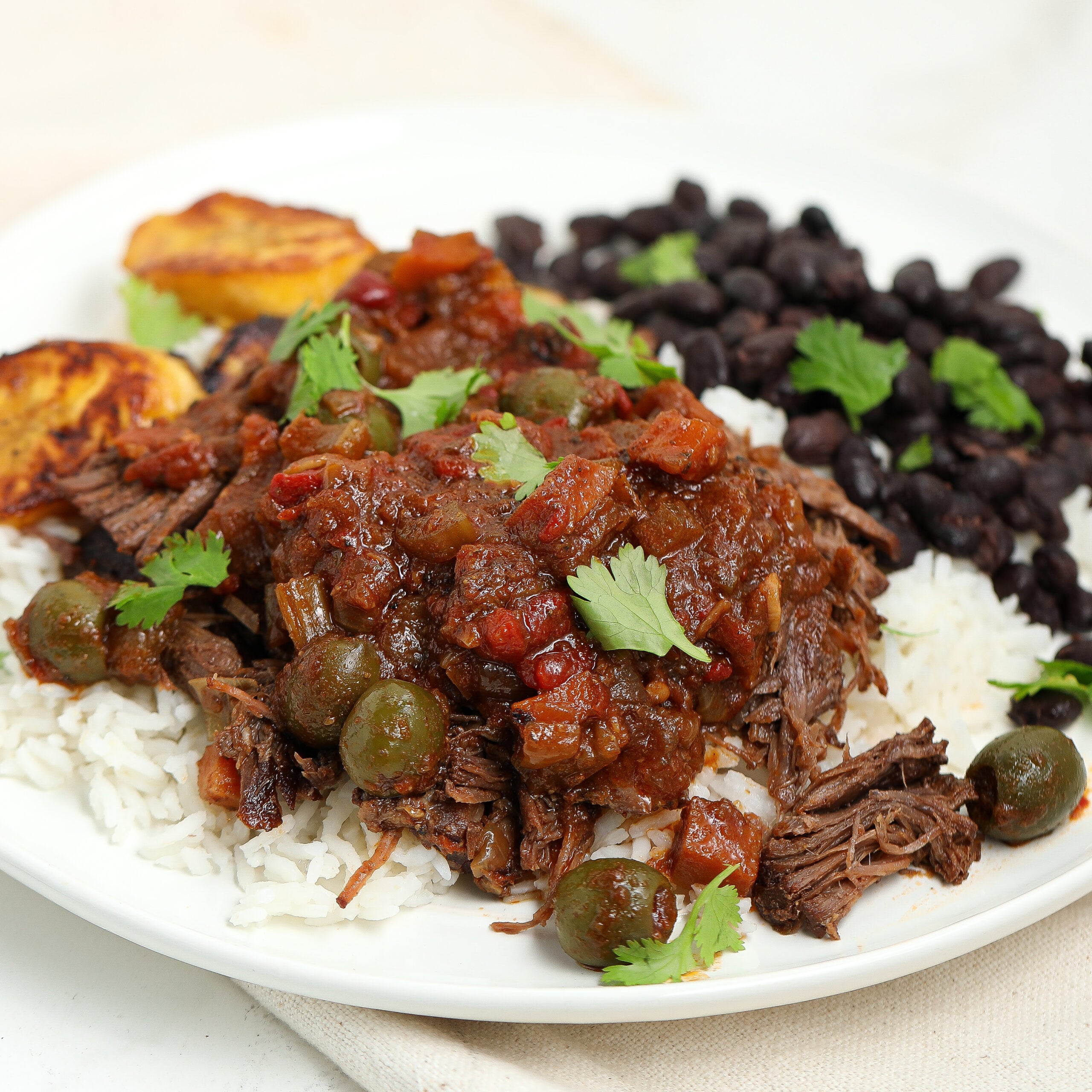 A plate of shredded beef in tomato sauce with olives served over white rice, accompanied by black beans garnished with cilantro and fried plantains.