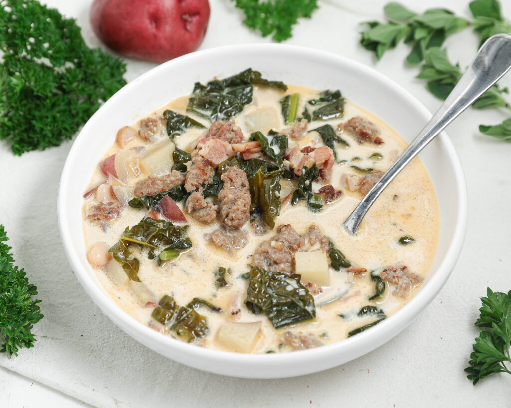 Creamy Italian sausage soup with kale, red potatoes, pancetta, and white beans in a white bowl with a spoon.