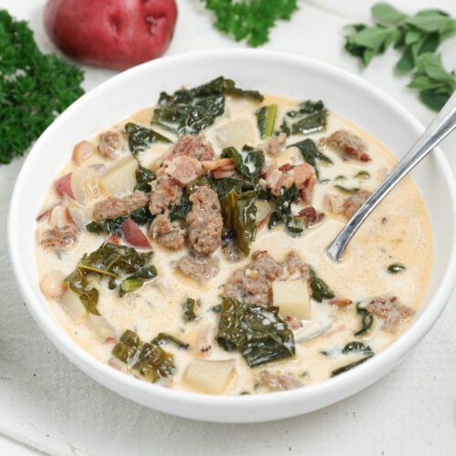 Creamy Italian sausage soup with kale, red potatoes, pancetta, and white beans in a white bowl with a spoon.