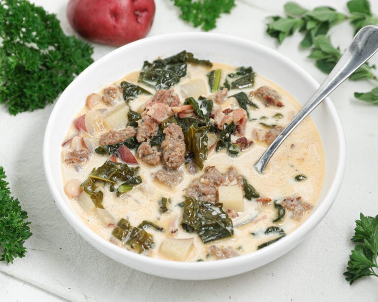 Creamy Italian sausage soup with kale, red potatoes, pancetta, and white beans in a white bowl with a spoon.