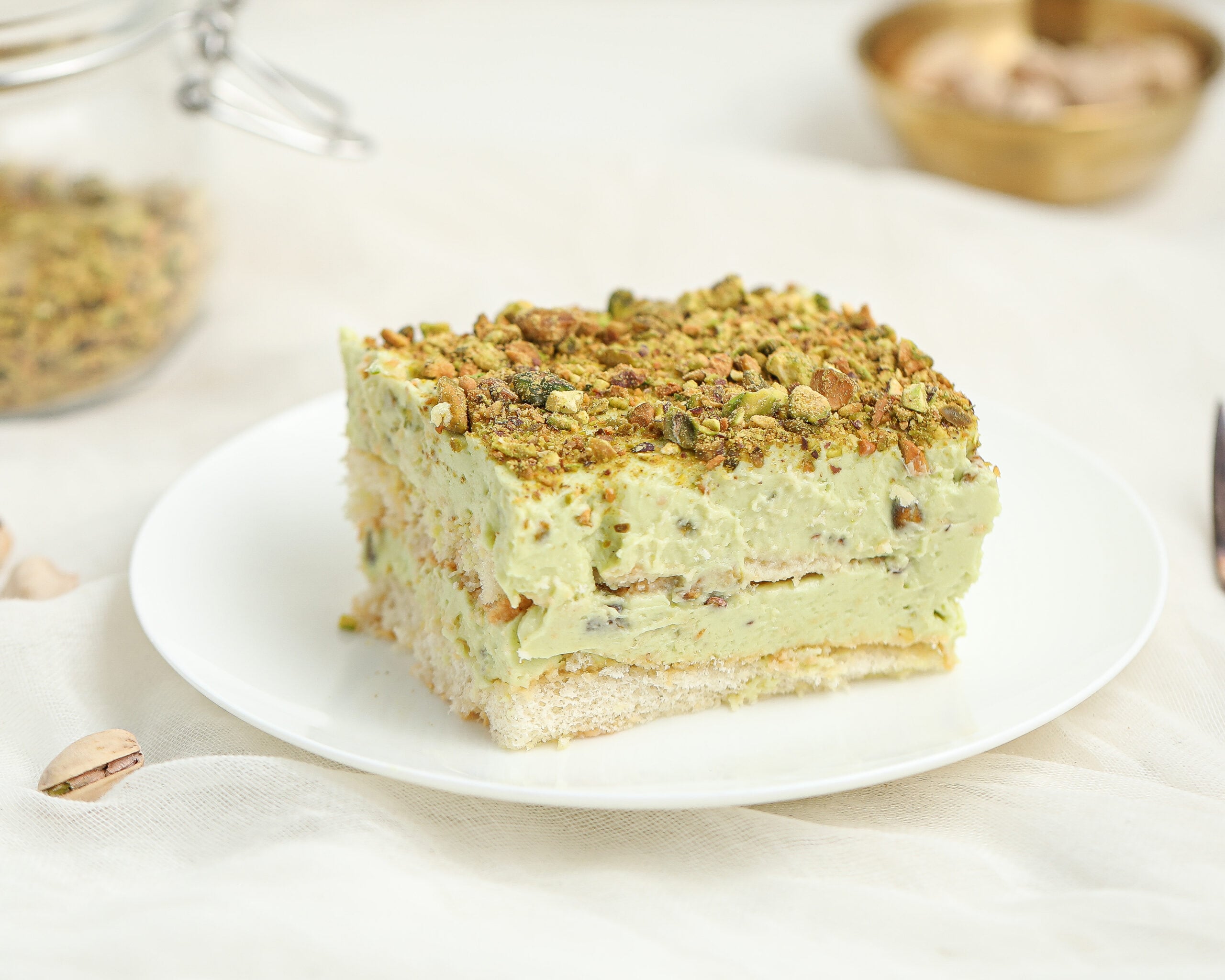 A slice of pistachio dessert with creamy green layers and a crumbly crust, topped with chopped pistachios, served on a white plate against a light background.