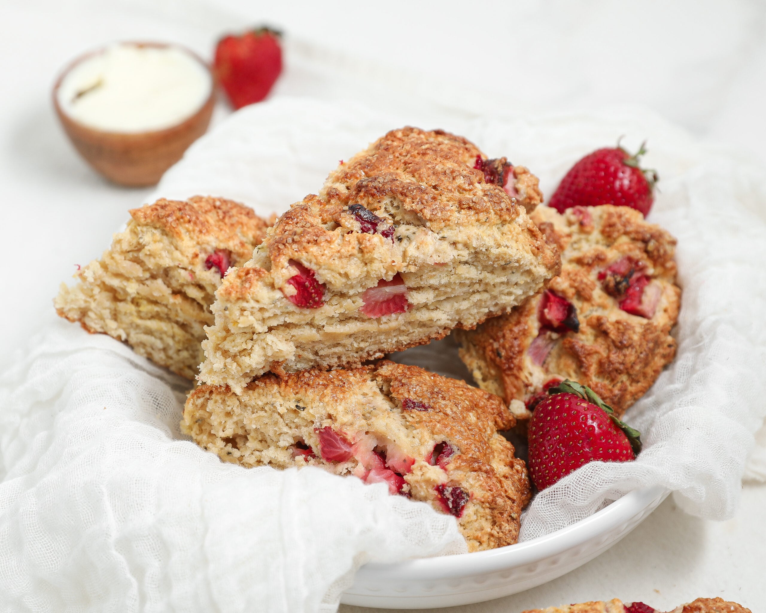 Strawberry basil scones stacked in a white bowl, showing flaky layers and fresh strawberry pieces, with whole strawberries and a small bowl of glaze in the background.
