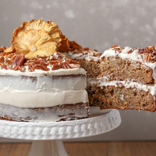 Hummingbird cake with cream cheese frosting topped with chopped pecans and dried pineapple, with a slice lifted from the cake stand.