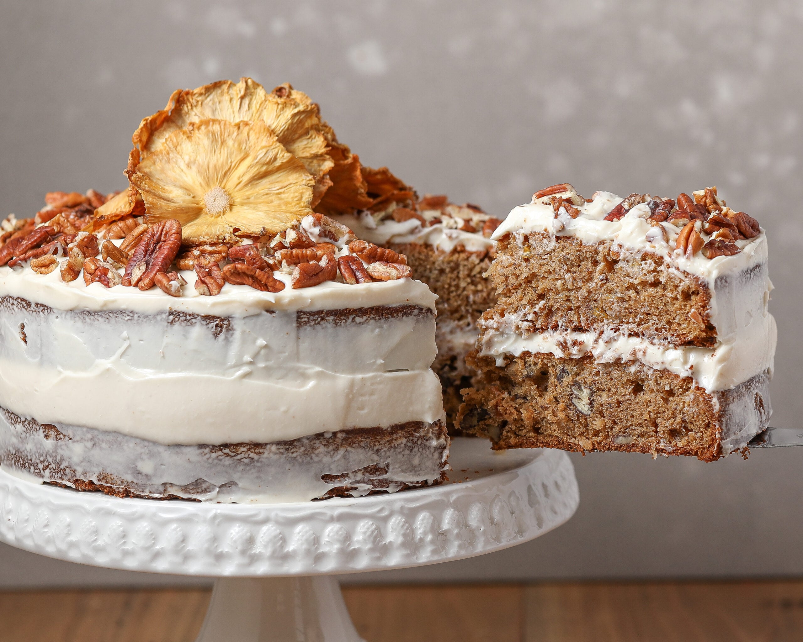 Hummingbird cake with cream cheese frosting topped with chopped pecans and dried pineapple, with a slice lifted from the cake stand.