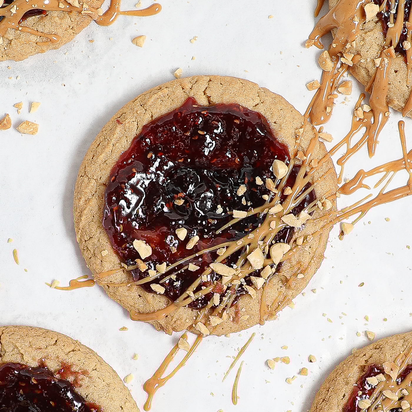 A gluten free almond butter and jelly cookie topped with a layer of dark red jam, drizzled with almond butter, and sprinkled with chopped peanuts, shown on a white background.