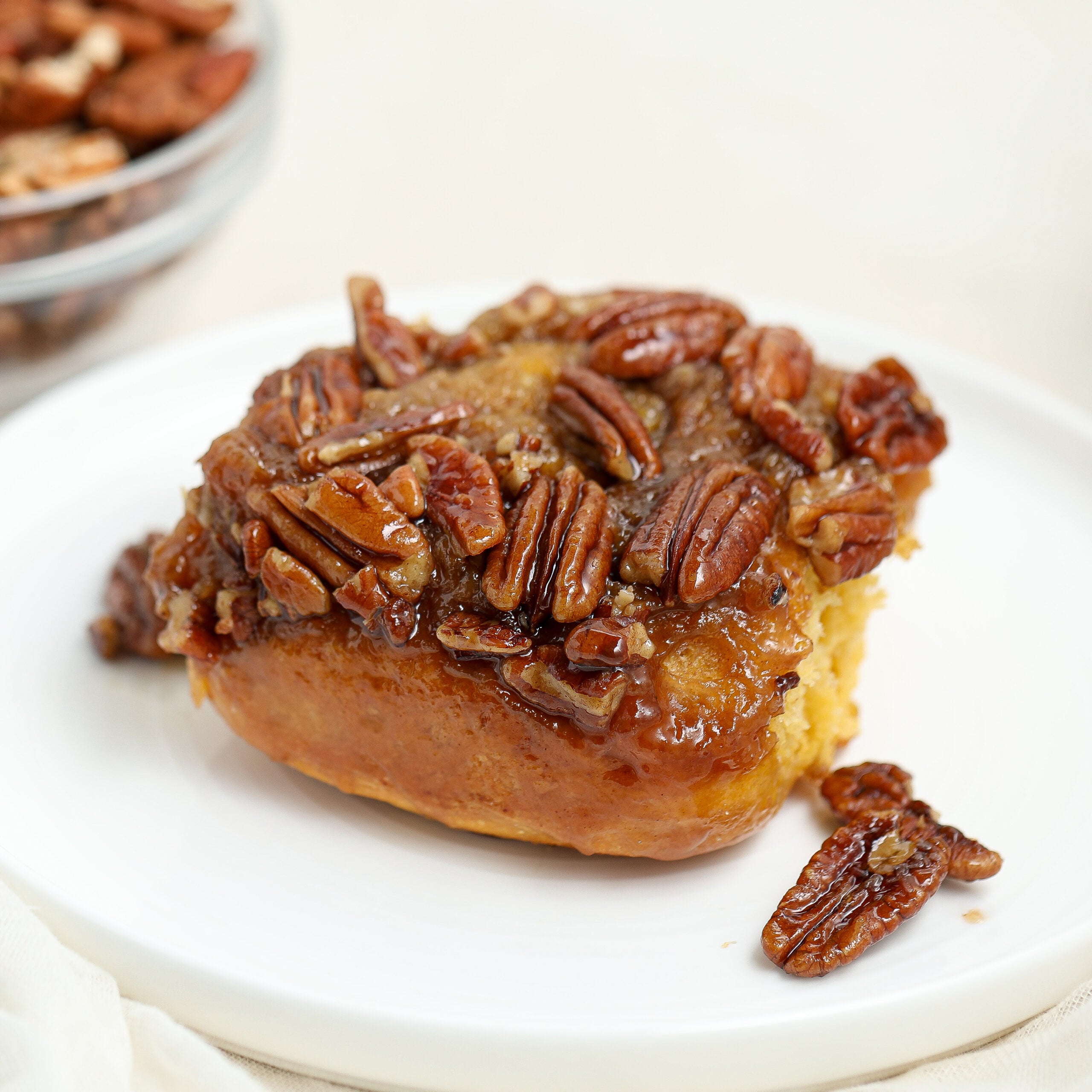 A sticky sweet potato bun topped with glazed pecans sits on a white plate, with a bowl of pecans blurred in the background.