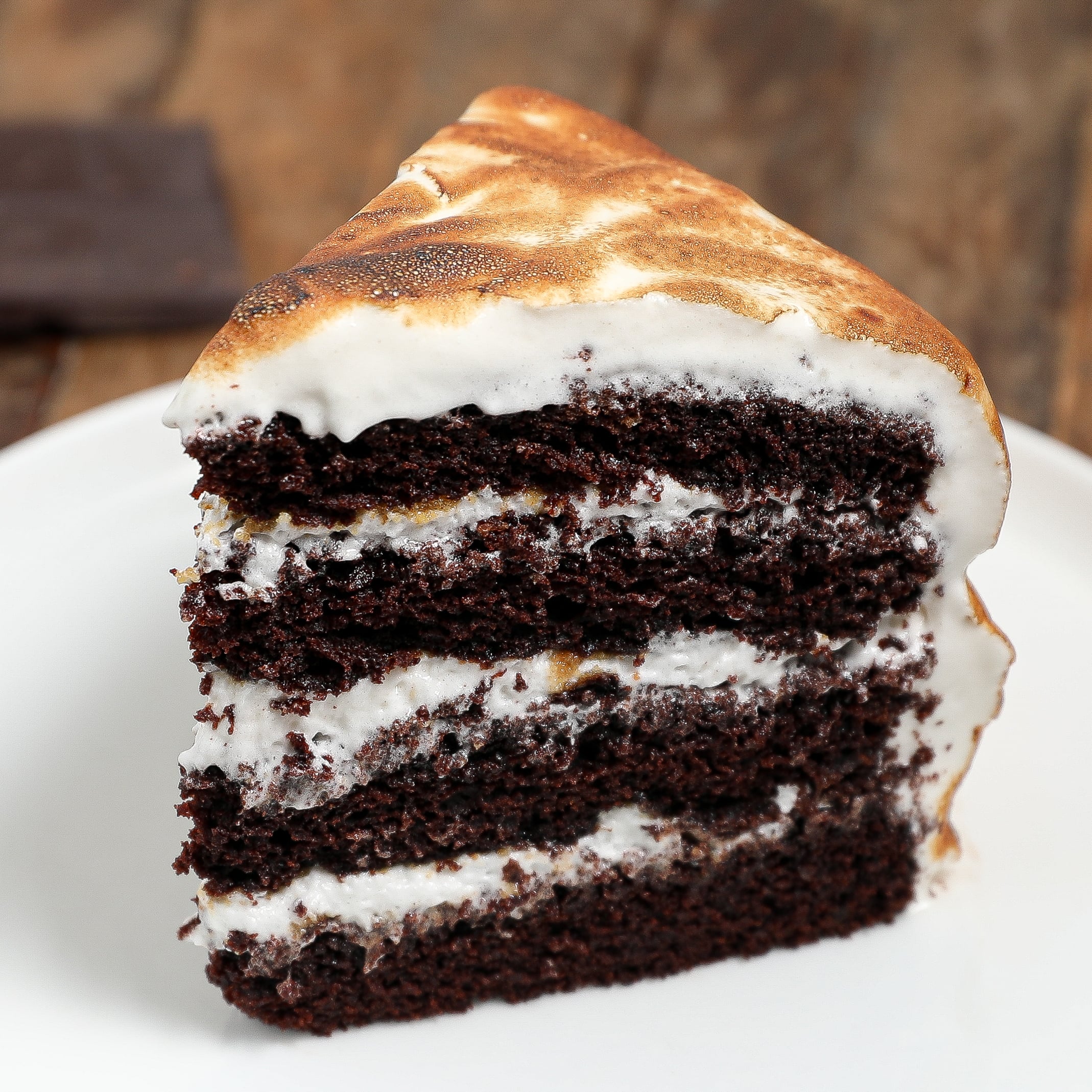A slice of decadent s'mores layer cake with white frosting between each layer and on top, served on a white plate against a rustic wooden background.