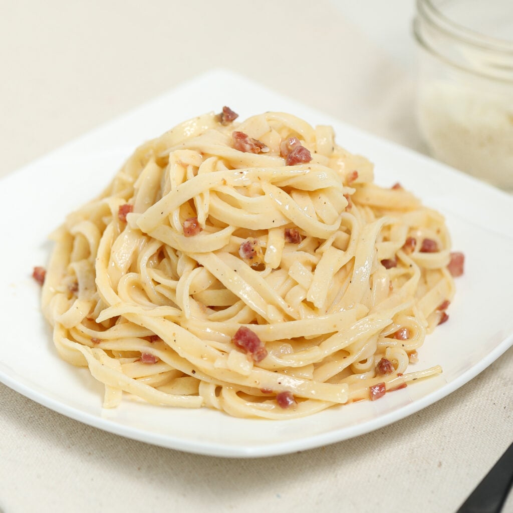A plate of fettuccine pasta with small pieces of bacon or pancetta, lightly coated in a creamy sauce, served on a white square dish with a jar in the blurred background.