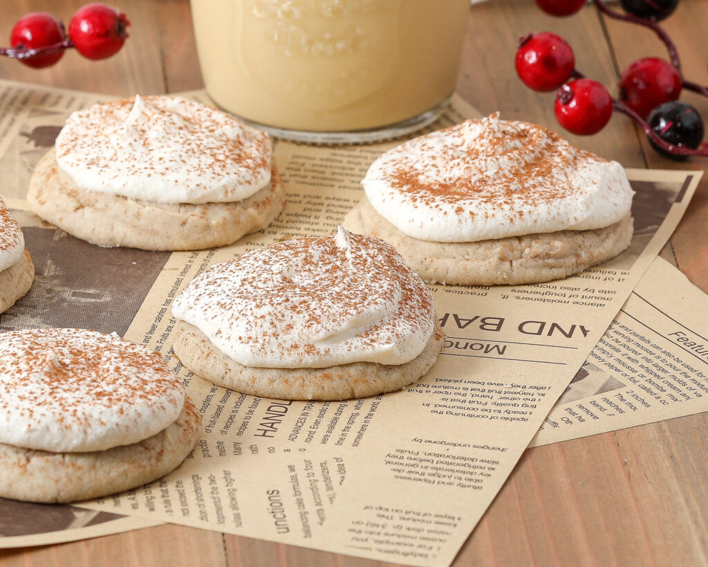 Four cookies topped with whipped cream and sprinkled with cinnamon sit on newspaper sheets atop a wooden surface. Red berries and a jar of a creamy drink are in the background.