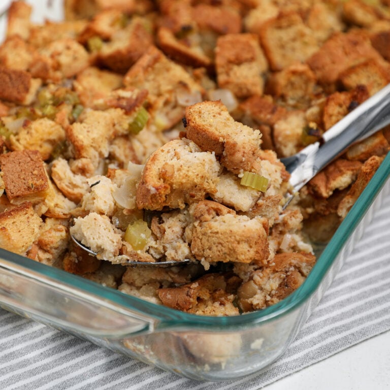 A glass baking dish filled with golden-brown herb bread stuffing mixed with celery and onions. A serving spoon is scooping out a portion as the dish rests on a gray and white striped cloth.