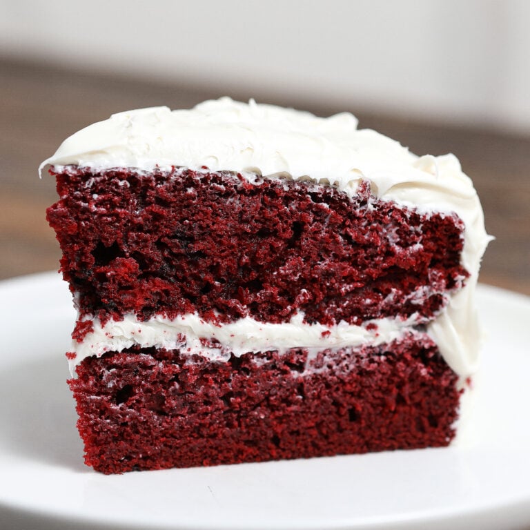 Close-up slice of homemade red velvet cake with a rich cream cheese frosting, showing a moist, velvety crumb and thick frosting layers on a white plate.
