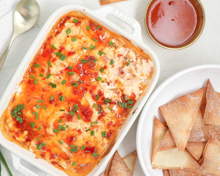 Creamy crab rangoon dip baked in a white dish, topped with chives and sweet chili sauce, served with crispy wonton chips.