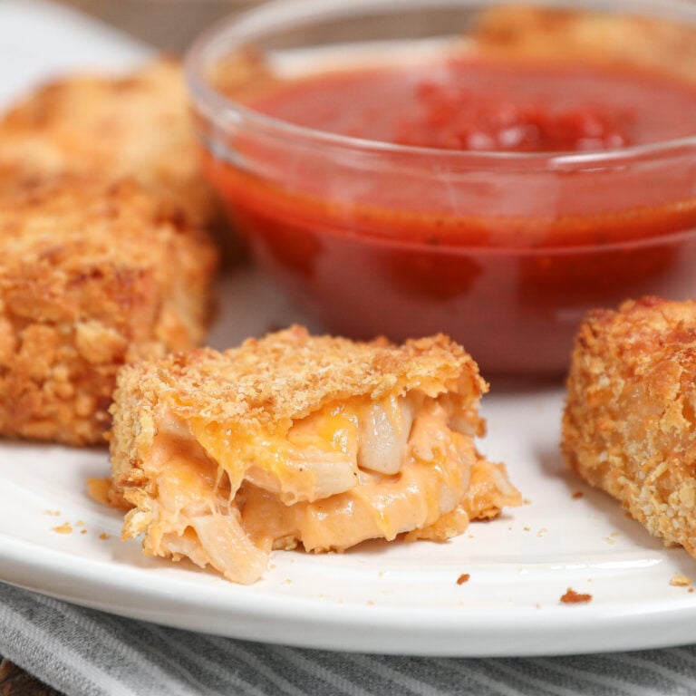 A close-up of crispy Gluten and Dairy Free Fried Macaroni and Cheese Bites on a white plate, with one bite showing gooey melted cheese and onions inside. A glass bowl of red dipping sauce sits in the background.