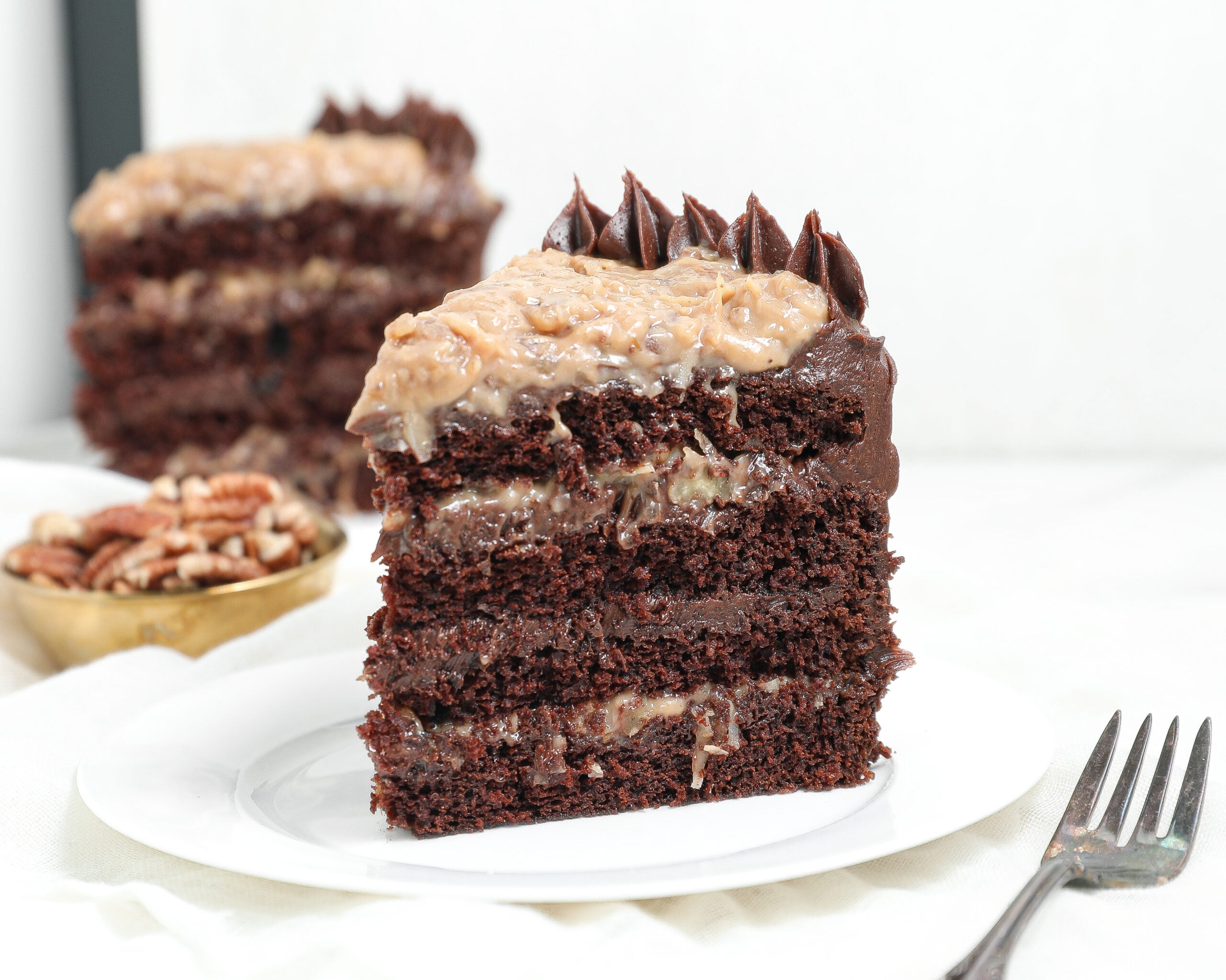 A slice of German chocolate cake with chocolate layers and coconut-pecan frosting sits on a white plate. A fork is beside the plate, and a bowl of pecans and another cake slice are in the background.