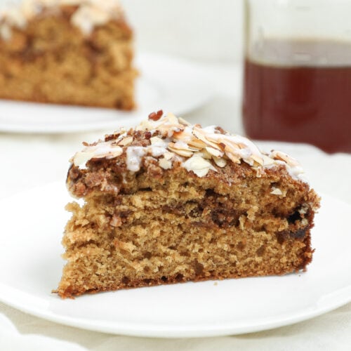 A slice of Honey Banana Coffee Cake topped with sliced almonds and glaze sits on a white plate. In the background, there is another slice of cake and a glass of dark liquid.