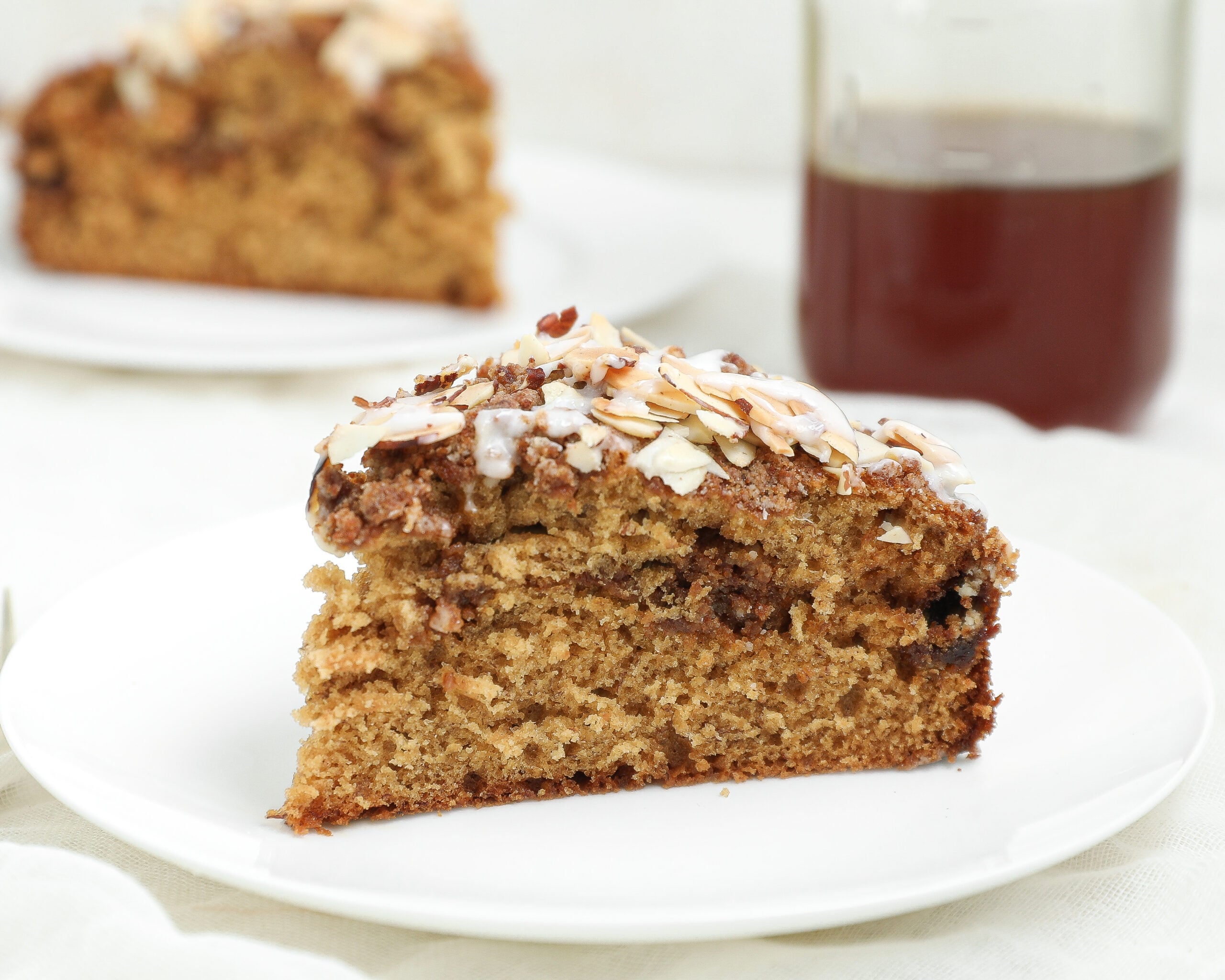 A slice of Honey Banana Coffee Cake topped with sliced almonds and glaze sits on a white plate. In the background, there is another slice of cake and a glass of dark liquid.