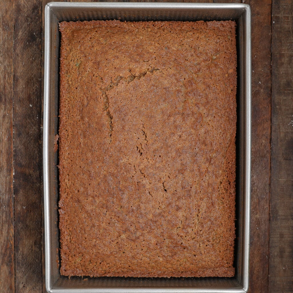 A rectangular, golden-brown Zucchini Carrot Cake sits in a metal baking pan on a wooden surface. The cake has a slightly cracked top and a textured, even surface.