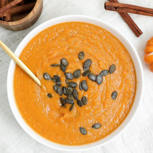 Creamy roasted butternut squash soup topped with pumpkin seeds in a white bowl with a gold spoon.