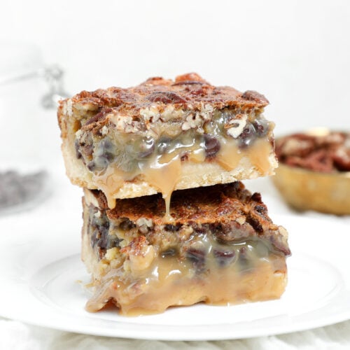 Two stacked chocolate pecan pie bars with a buttery shortbread crust, pecan topping, and gooey caramel-like filling dripping down the sides.