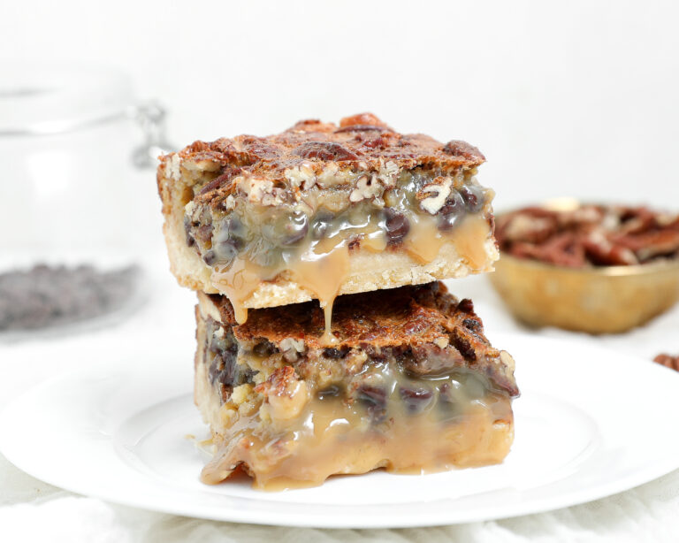 Two stacked chocolate pecan pie bars with a buttery shortbread crust, pecan topping, and gooey caramel-like filling dripping down the sides.