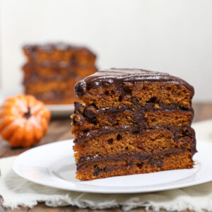 A slice of Pumpkin Cake with Chocolate Chips, topped with rich chocolate frosting, sits on a white plate. Another slice and a small decorative pumpkin appear in the blurred background, highlighting the cake’s moist layers and indulgent chocolate filling.