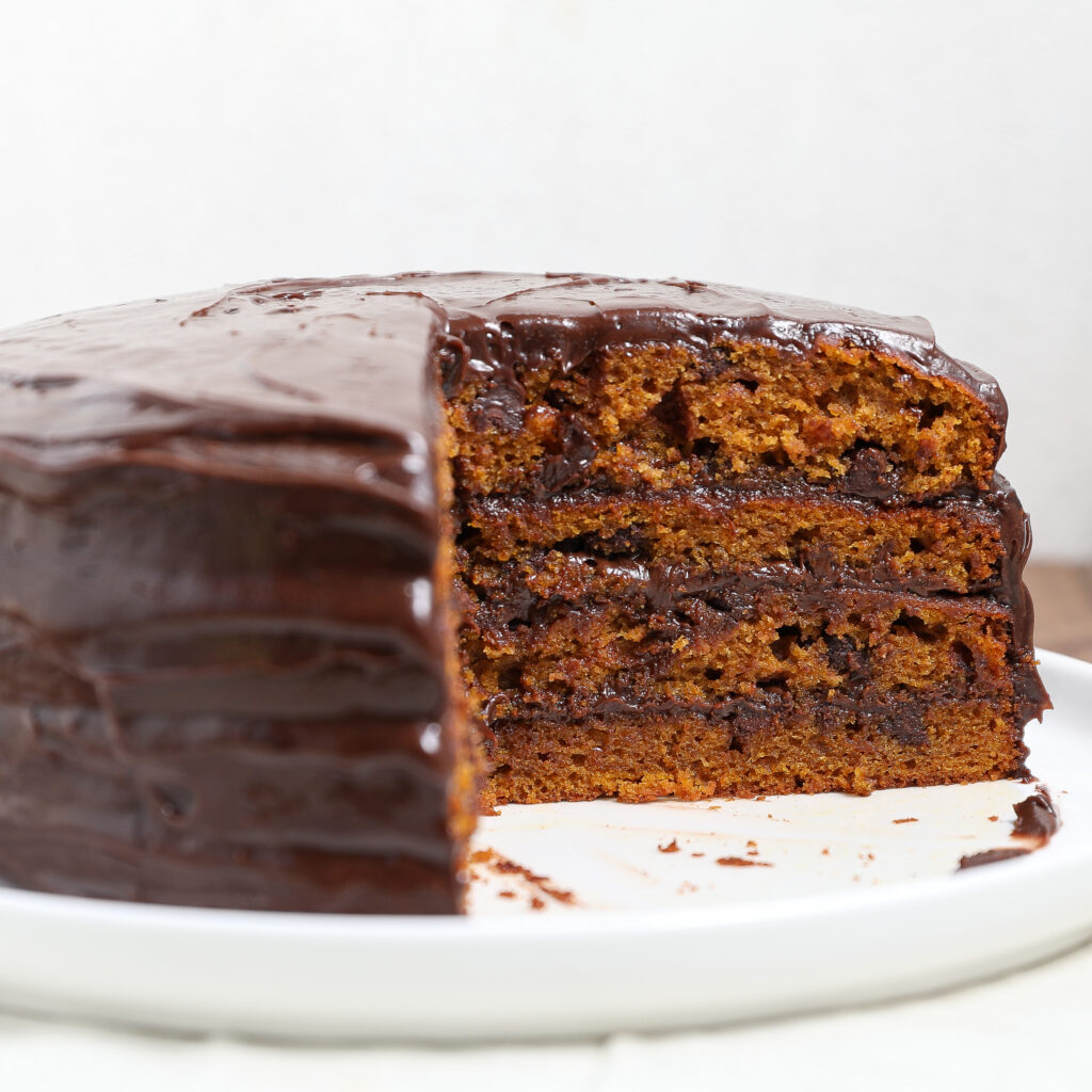 A three-layer Pumpkin Cake with Chocolate Chips and chocolate frosting sits on a white plate. A large slice has been removed, revealing the moist, rich interior filled with chocolate chips.