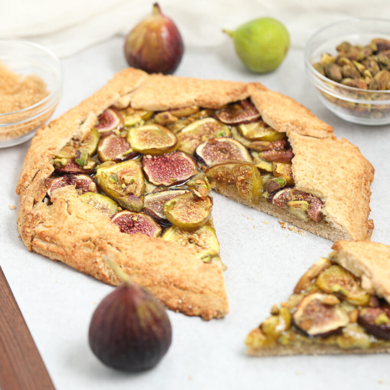 A rustic fig galette with a slice cut out, topped with fresh figs and nuts, sits on parchment paper. Whole figs, a bowl of brown sugar, and a bowl of pistachios are placed around the galette.