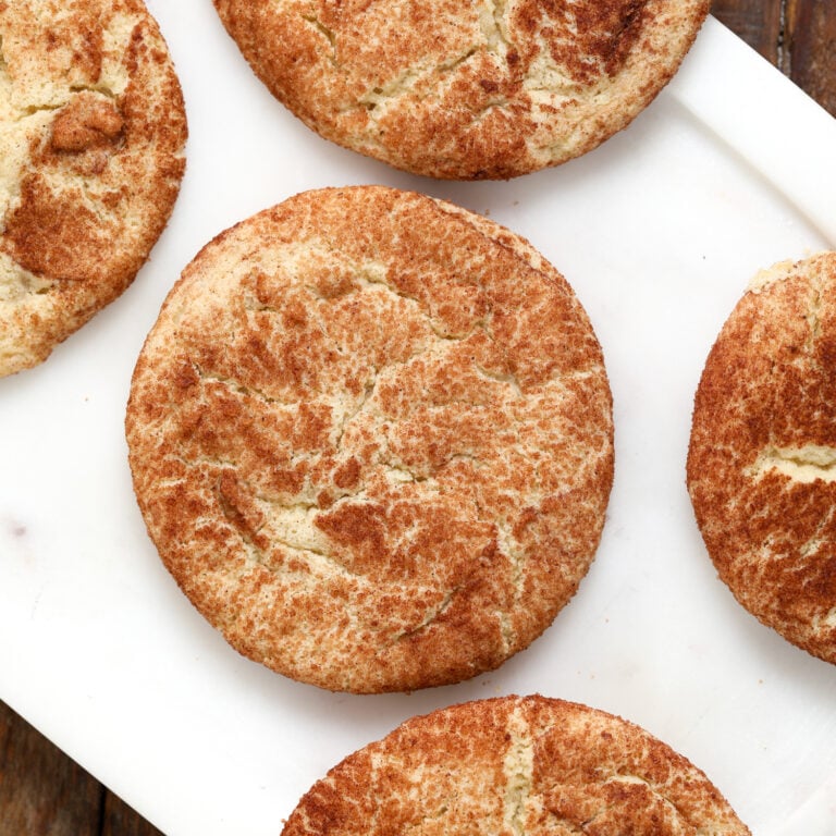 Five golden-brown Snickerdoodle Cookies with crackled tops are arranged on a white rectangular surface, some partially cropped at the edges.