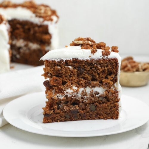 Slice of layered carrot cake with cream cheese frosting on a white plate, topped with walnuts and cake crumbs.