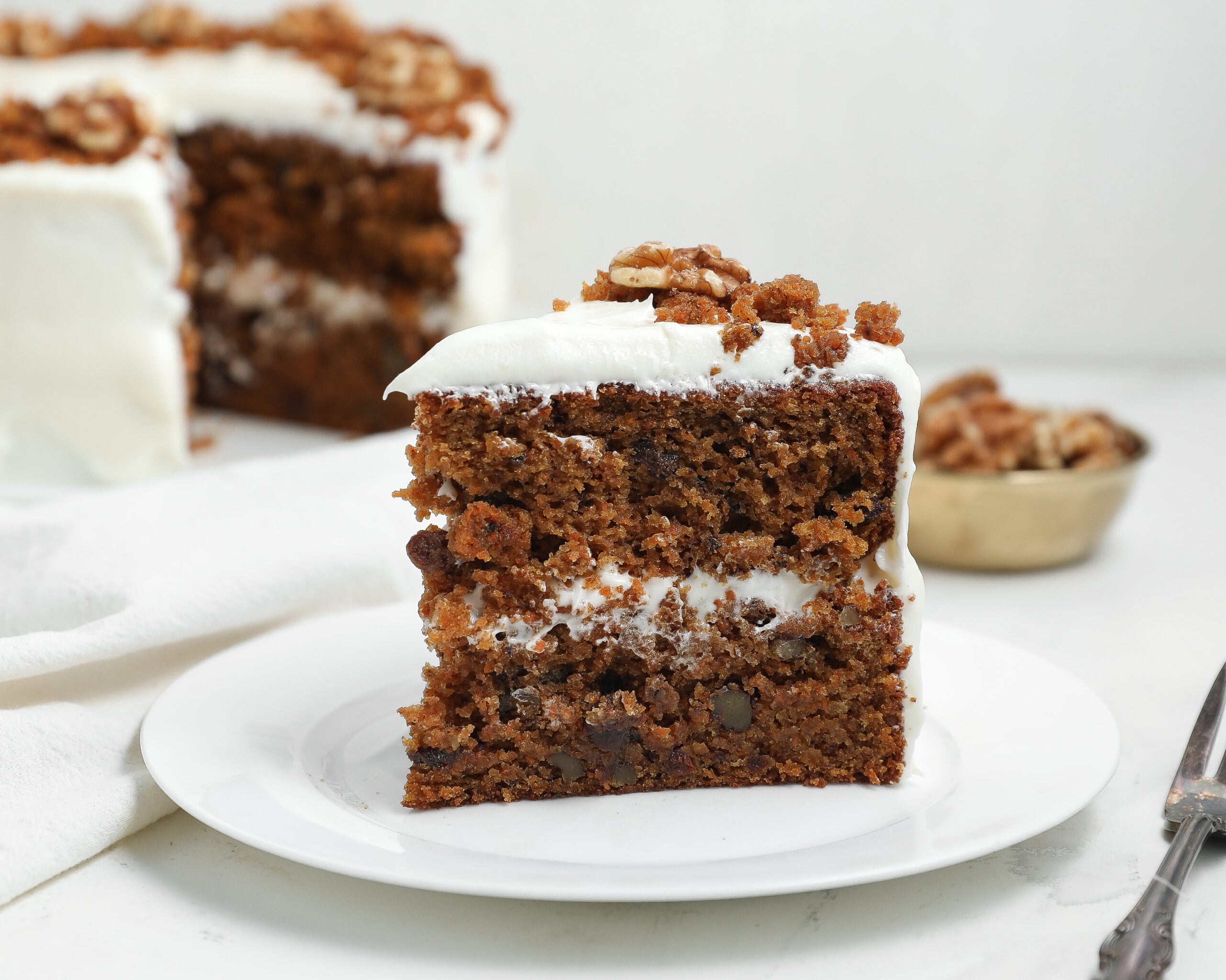 Slice of layered carrot cake with cream cheese frosting on a white plate, topped with walnuts and cake crumbs.