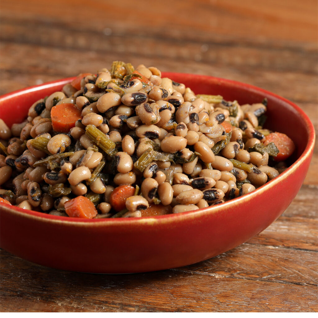 A red bowl filled with Easy Smoky Black-Eyed Peas, mixed with chopped carrots and greens, sits on a wooden surface.