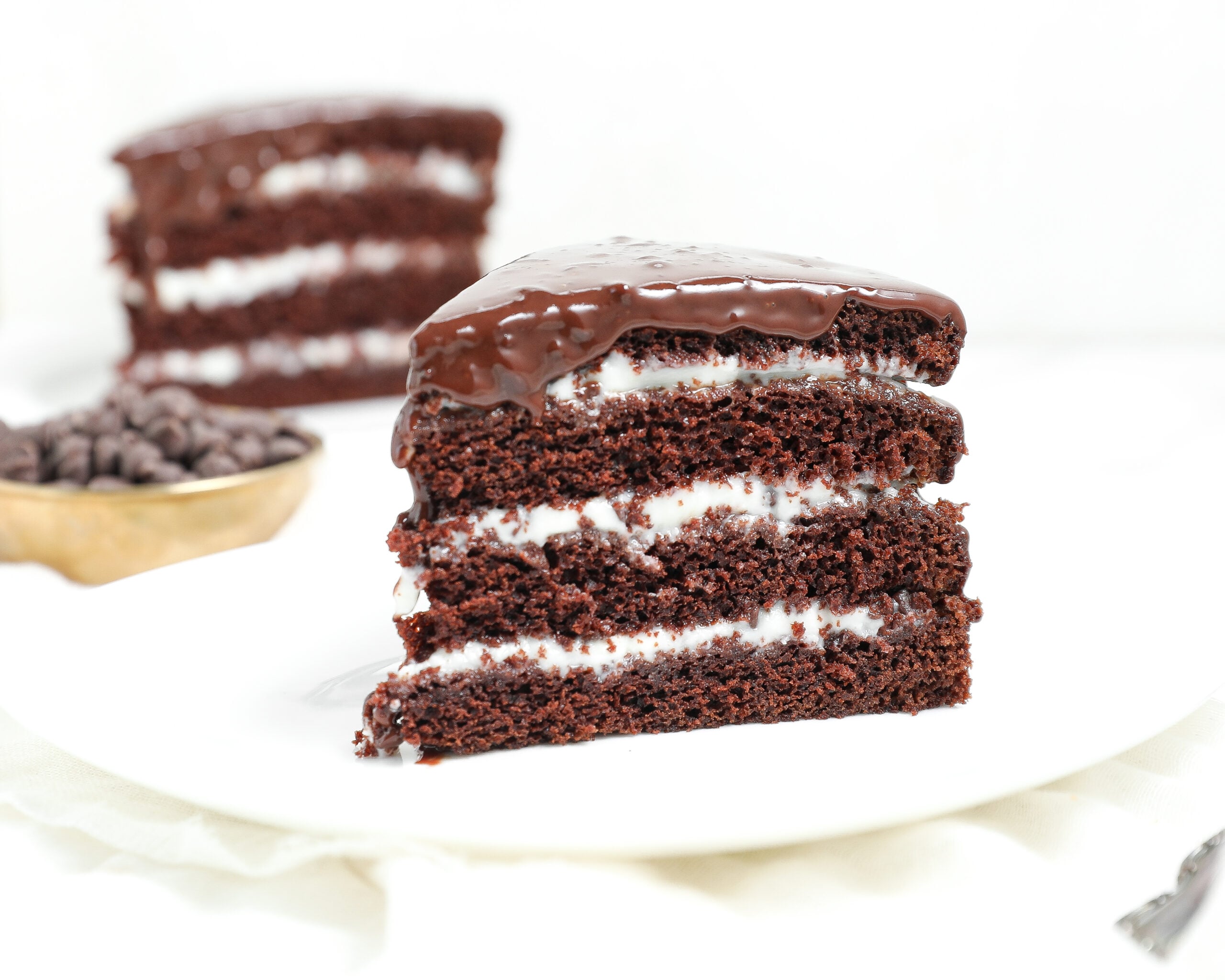 A slice of four-layer chocolate cake with white cream filling and glossy chocolate glaze on top is displayed on a white plate, with another slice and a bowl of chocolate chips in the background.