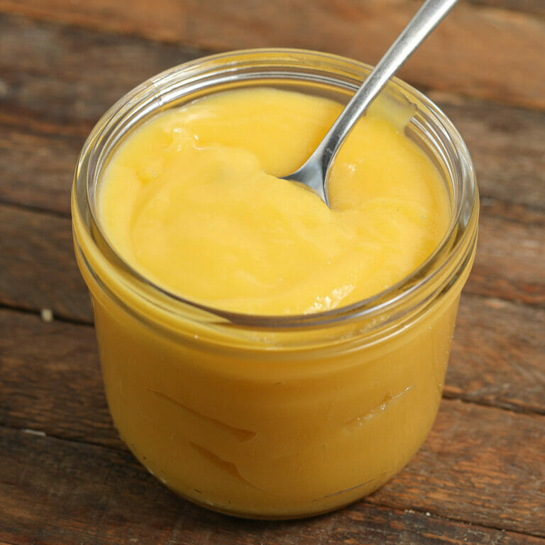 A glass jar filled with smooth, yellow passion fruit curd sits on a wooden surface, with a metal spoon resting inside the jar.