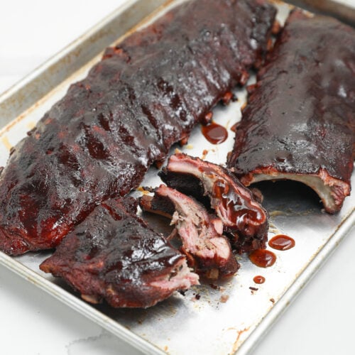 Two racks of barbecued ribs coated in dark sauce on a metal baking sheet, with one rack partially sliced to show tender, juicy meat inside.