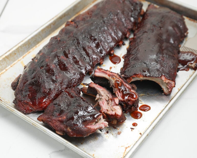 Two racks of barbecued ribs coated in dark sauce on a metal baking sheet, with one rack partially sliced to show tender, juicy meat inside.