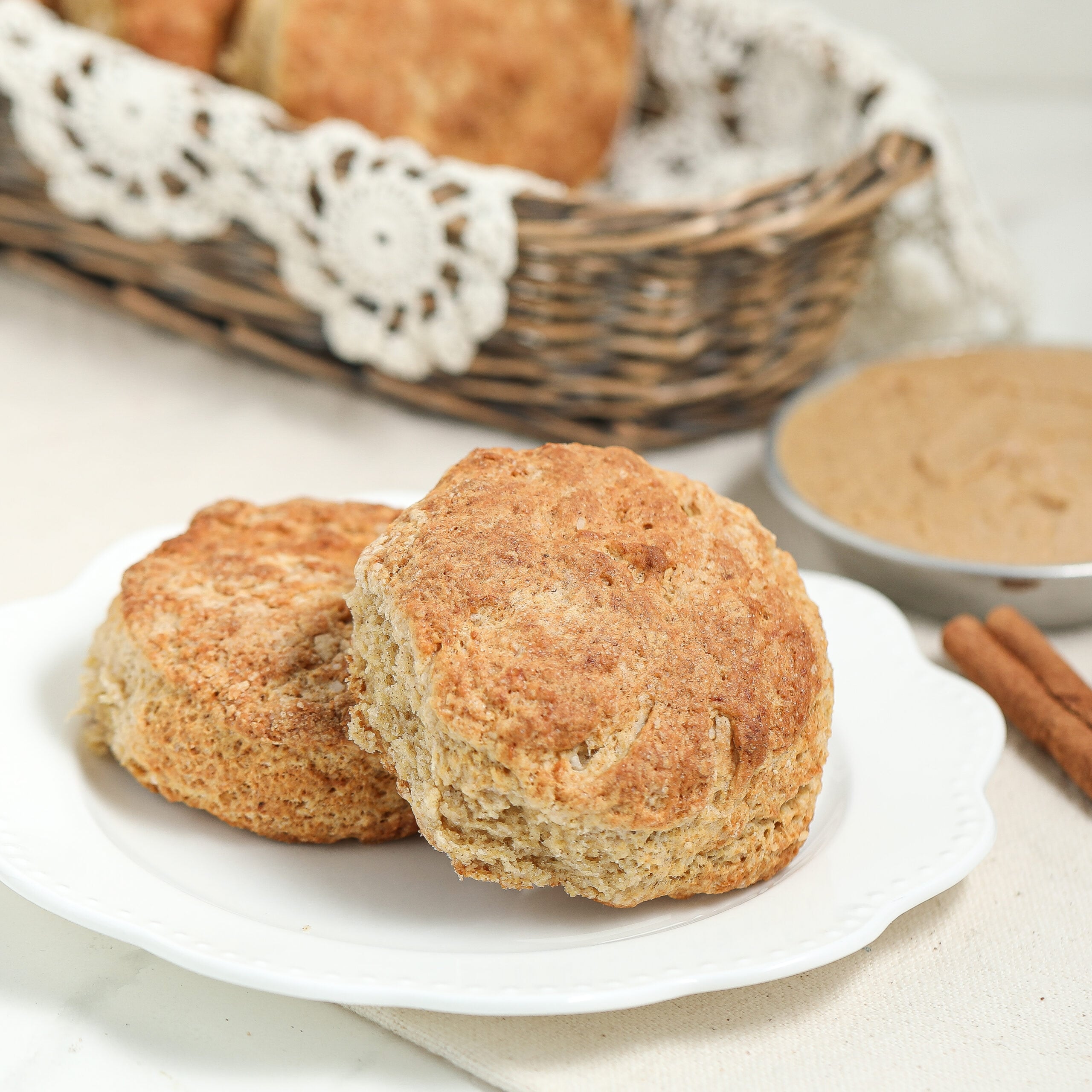 Two golden-brown biscuits on a white plate, with a wicker basket of more biscuits in the background and a small bowl of light brown spread nearby. A lace doily decorates the basket.