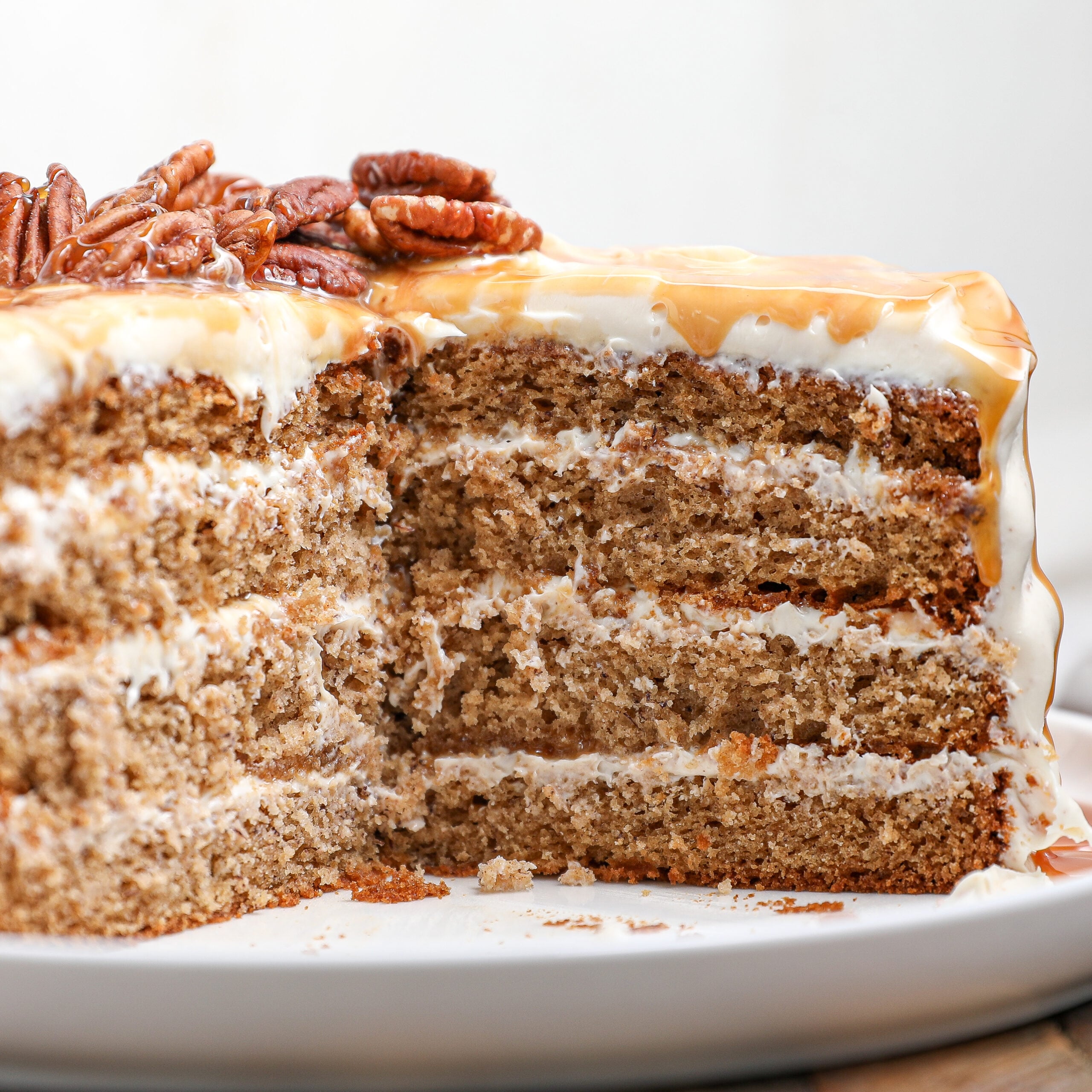 A close-up of a sliced banana cake with cream filling, topped with caramel glaze and pecans, on a white plate. The cake appears moist with visible layers and a creamy, nutty topping.