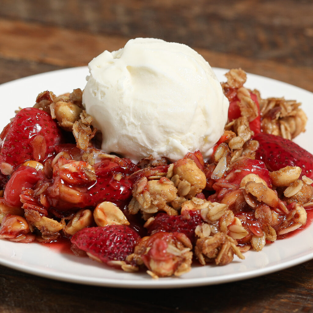 A plate of strawberry crisp topped with a scoop of vanilla ice cream. Reminiscent of blueberry peach cobbler, this dessert features baked strawberries and oats, with melting ice cream adding creamy contrast on a rustic wooden surface.