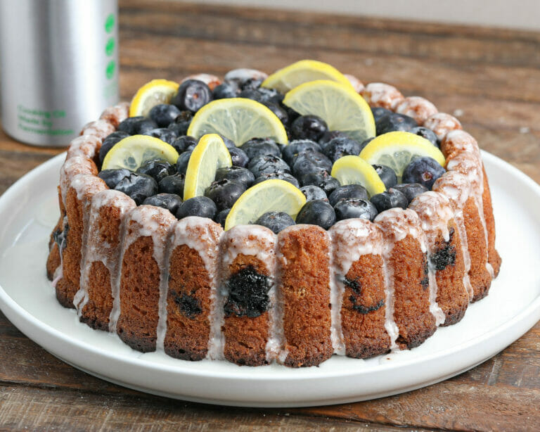 A Lemon Blueberry Cake with white icing drizzled on top, garnished with fresh blueberries and lemon slices, sits on a white plate on a rustic wooden table.