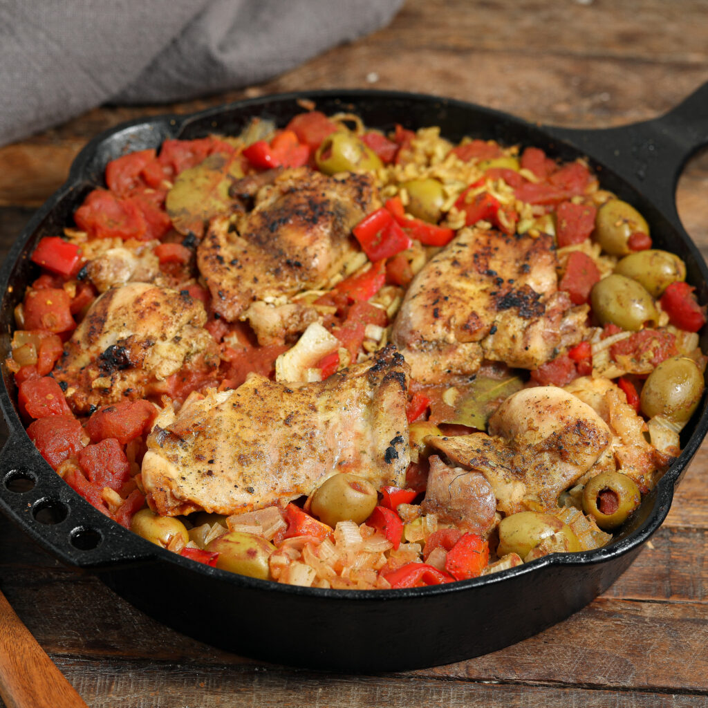 A cast iron skillet filled with cooked chicken thighs, green olives, diced tomatoes, onions, and red bell peppers rests on a wooden surface—a vibrant mix reminiscent of classic arroz con pollo.