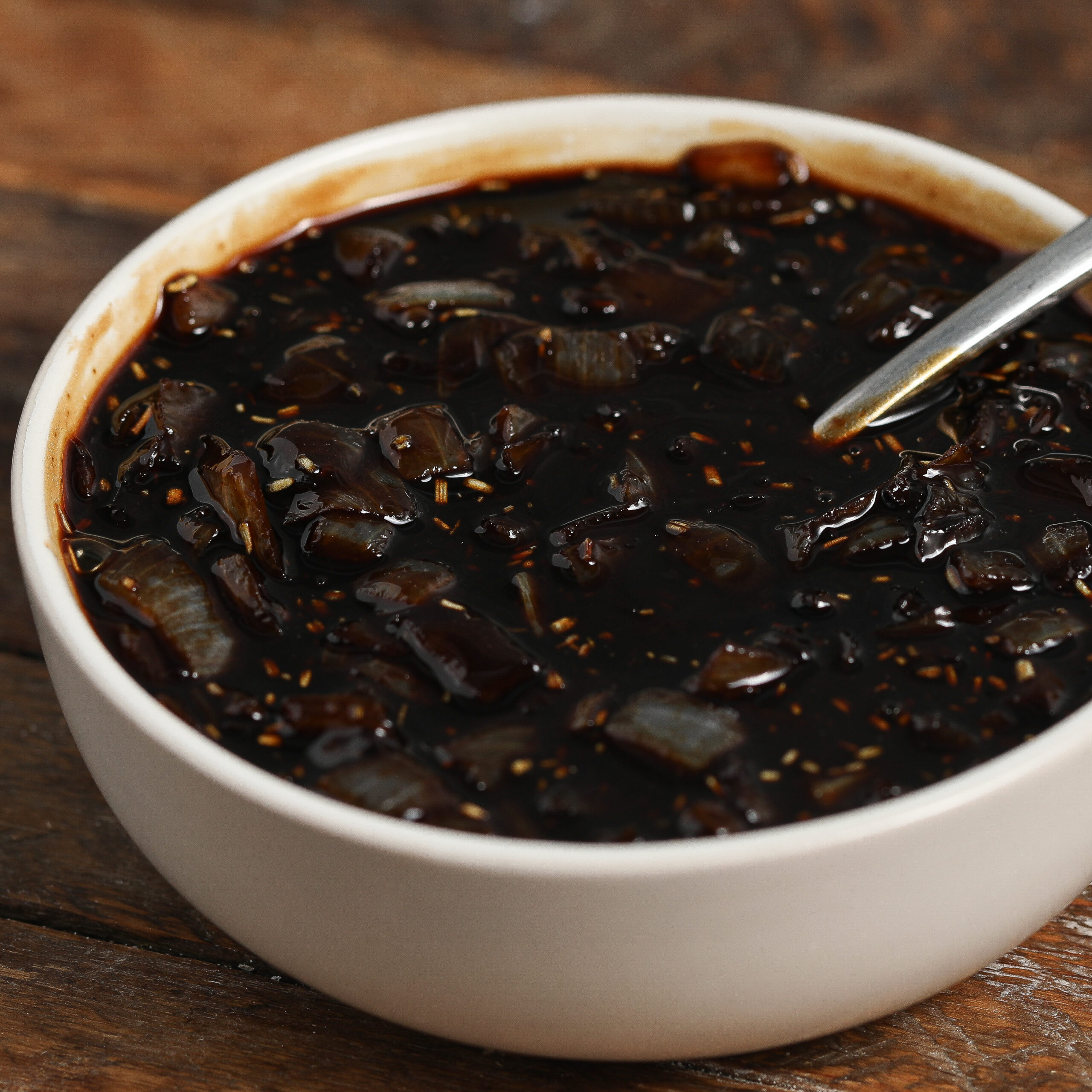 A white bowl filled with dark, chunky onion jam containing diced onions and spices, with a metal spoon resting inside, placed on a rustic wooden surface.