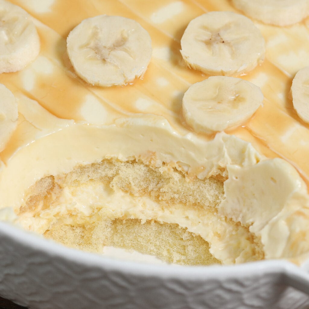 Close-up of a Banana Pudding Tiramisu in a white dish, topped with banana slices and drizzled caramel. A portion is scooped out, revealing layers of creamy pudding and sponge cake.