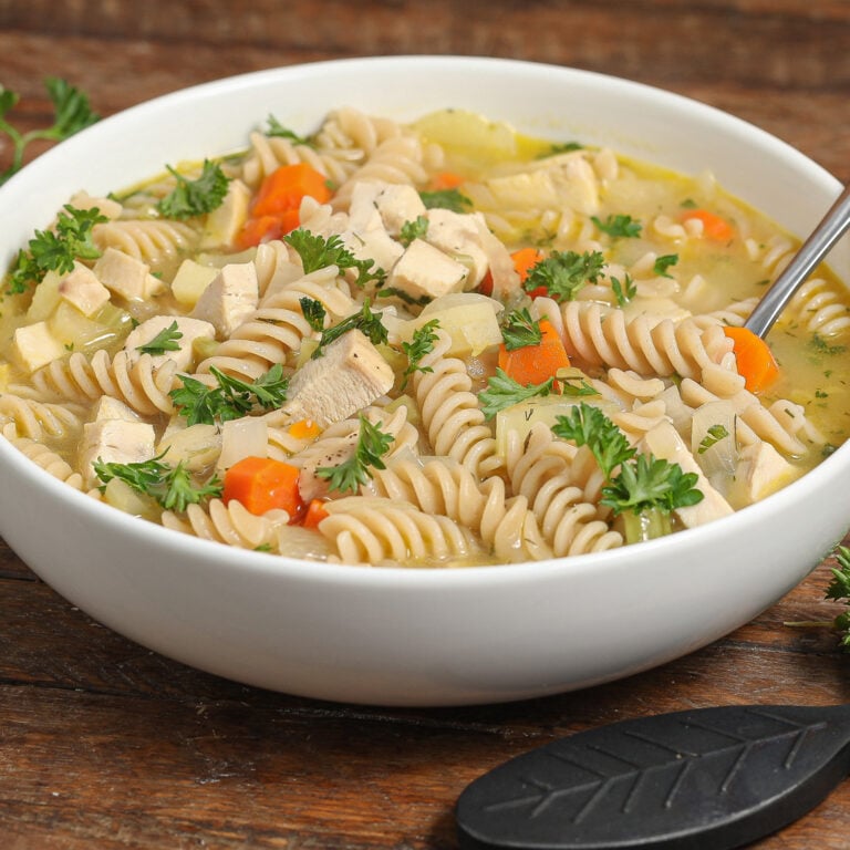 A white bowl filled with rotini chicken noodle soup, diced chicken, carrots, and fresh parsley garnish, with a spoon resting inside, on a wooden table.