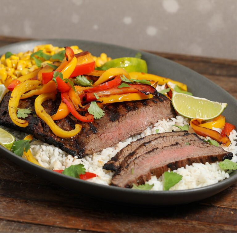 A flavorful Steak Fajita Rice Bowl featuring grilled steak slices topped with sautéed red and yellow bell peppers, served over white rice, garnished with cilantro, lime wedges, and a side of corn.