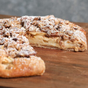 A rustic Apple Crisp Galette with Cashew Butter features a crumbly topping and powdered sugar, sliced on a wooden surface to reveal layers of tender apple filling inside.