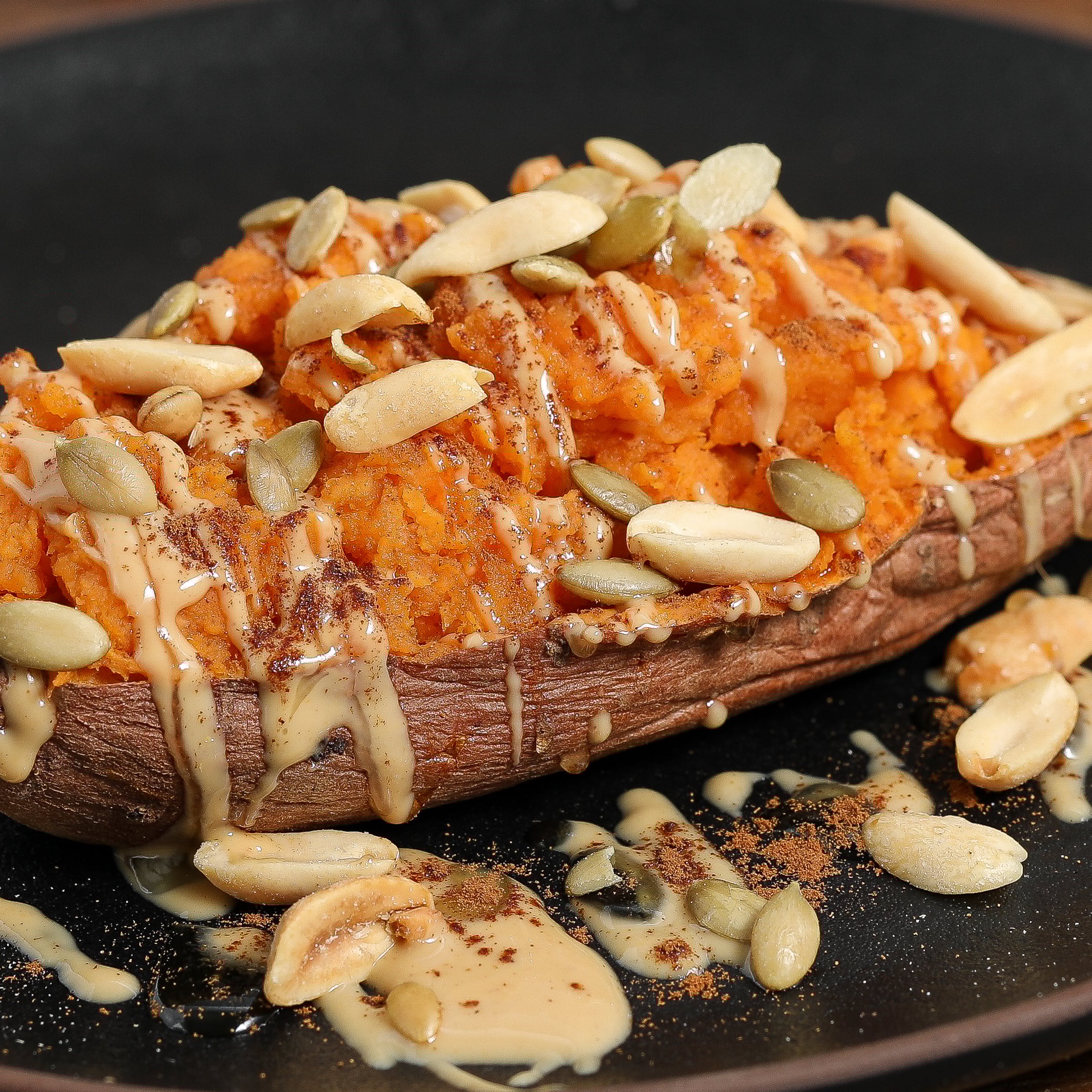 A loaded sweet potato with peanut butter sauce, topped with pumpkin seeds, peanuts, and spices, served on a black plate.