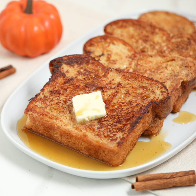 Three slices of French toast with syrup and a pat of butter on top are served on a white plate, with a small pumpkin and cinnamon sticks nearby.