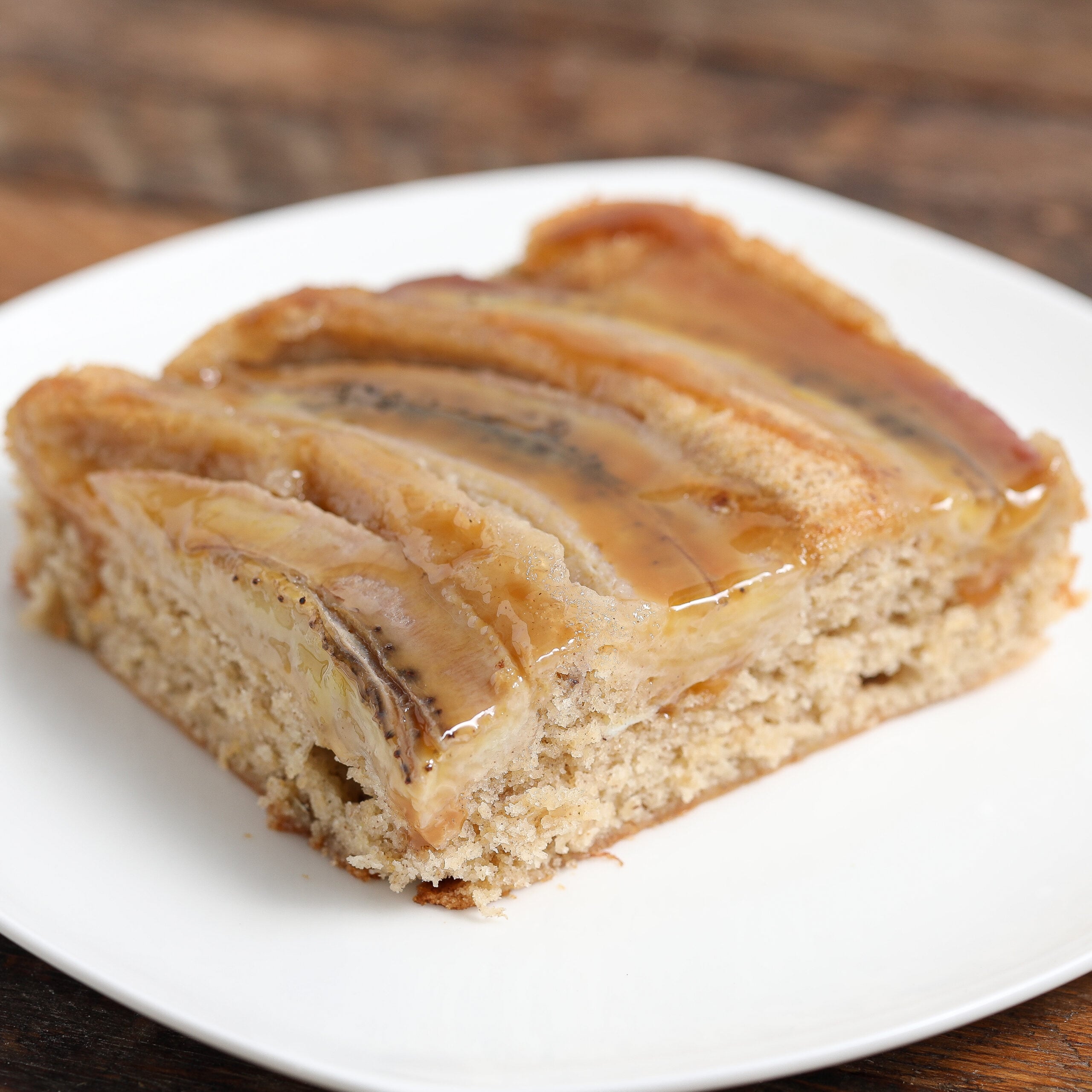 A square slice of gluten free banana caramel upside down cake sits on a white plate, topped with caramelized banana slices and a shiny glaze, with a moist, light brown cake base.
