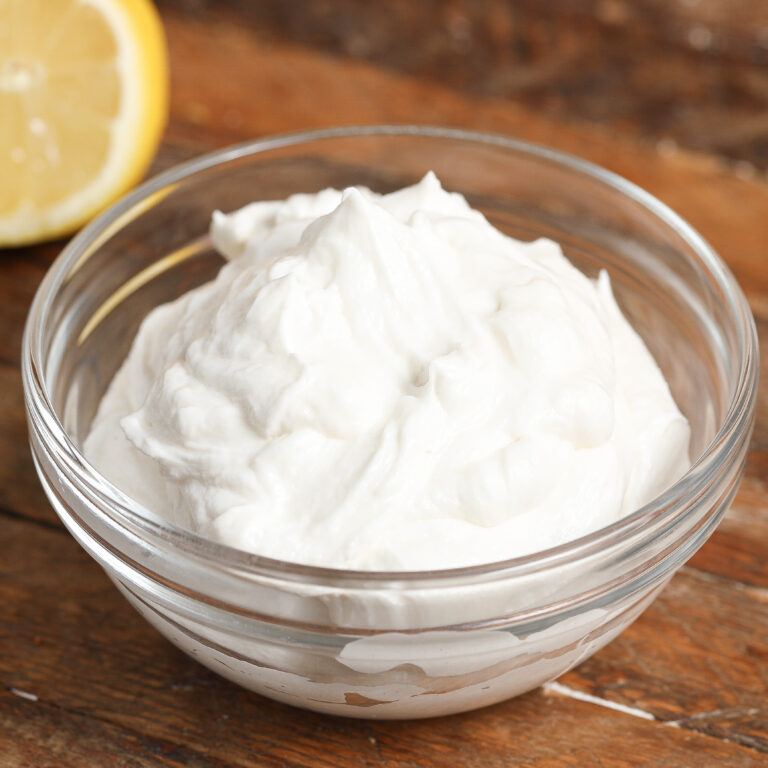 A clear glass bowl filled with thick, creamy white whipped feta sits on a wooden surface. A lemon wedge is partially visible in the background.
