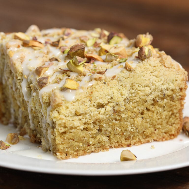 A close-up of a thick slice of pistachio crumb cake topped with white icing and chopped pistachios, served on a white plate against a wooden background.